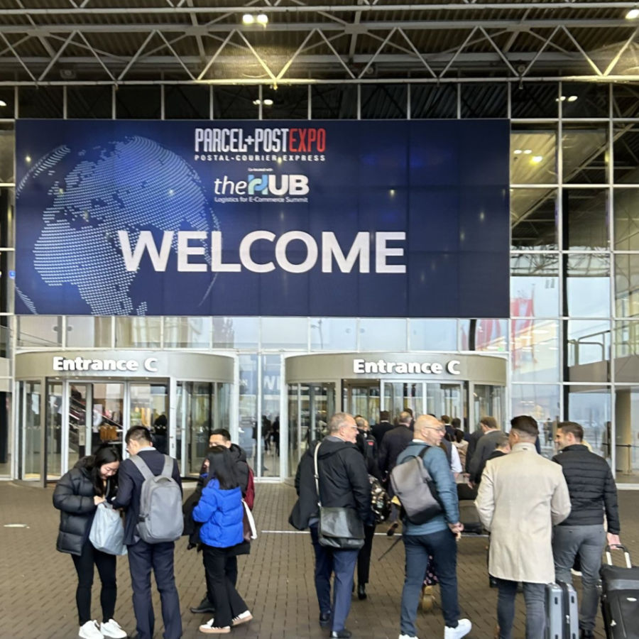 Eingang zur Ausstellung mit dem Schild - PARCEL+POST EXPO. Banner mit einer Weltkugel-Grafik. Türen - Entrance C. Gruppe von Menschen vor dem Eingang.