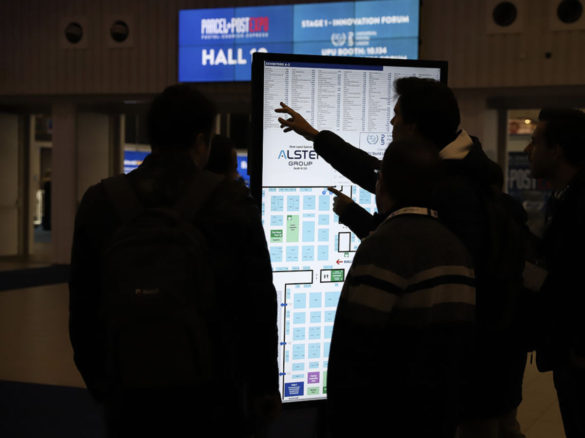 Vier Personen stehen vor einem Informationsdisplay. Einer zeigt auf den Messeplan. Im Hintergrund sind die Schilder - PARCEL+POST EXPO und - HALL C. Gedämpftes Licht.